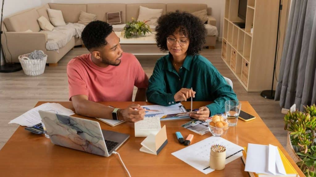 A serious couple sits at a desk, looking at documents and a laptop.