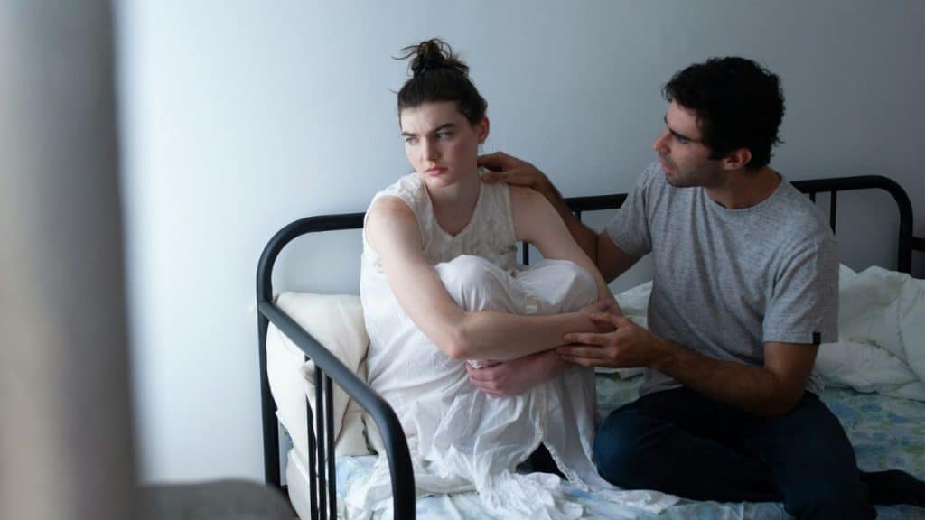 A worried man tries to comfort a sad, distraught woman on a bed.