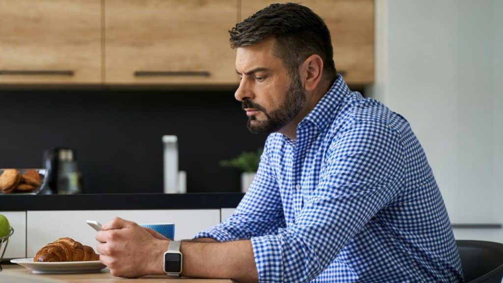 A focused man with a beard sits at a table, using his phone.