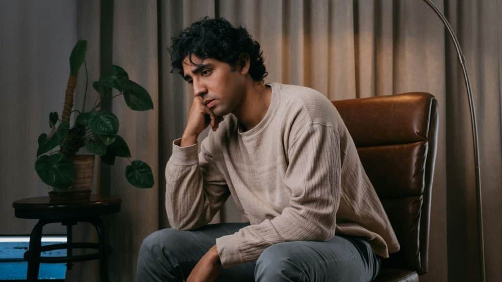 A man with dark, curly hair sits in a chair, looking sad.