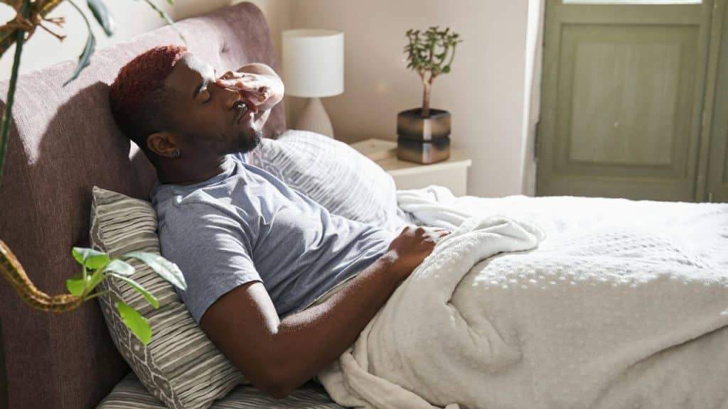 A sad man with red hair lies on his bed, covering his eyes with his hand.