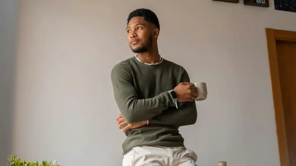 A man in a green sweater, holding a mug, looks away thoughtfully.