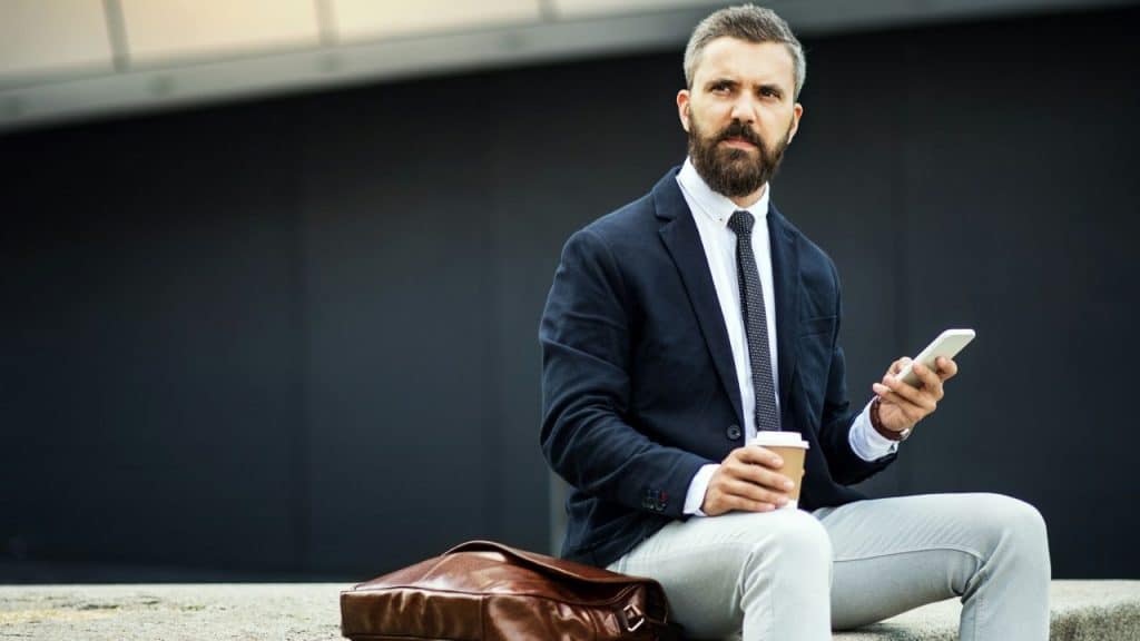 A stylish businessman with a beard holds a coffee and a phone, sitting outdoors.