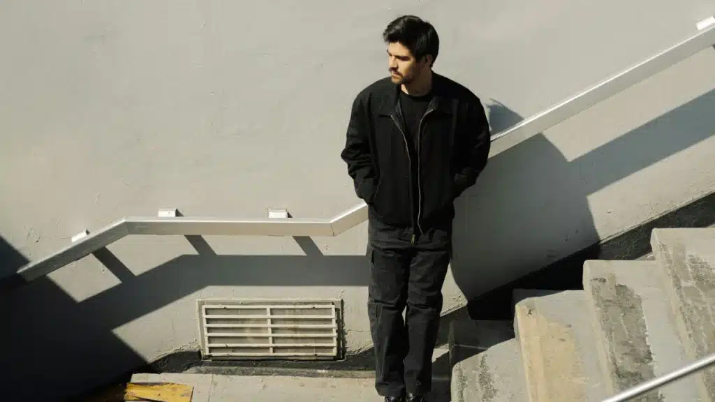 A man in a black jacket and cargo pants stands on a concrete staircase.