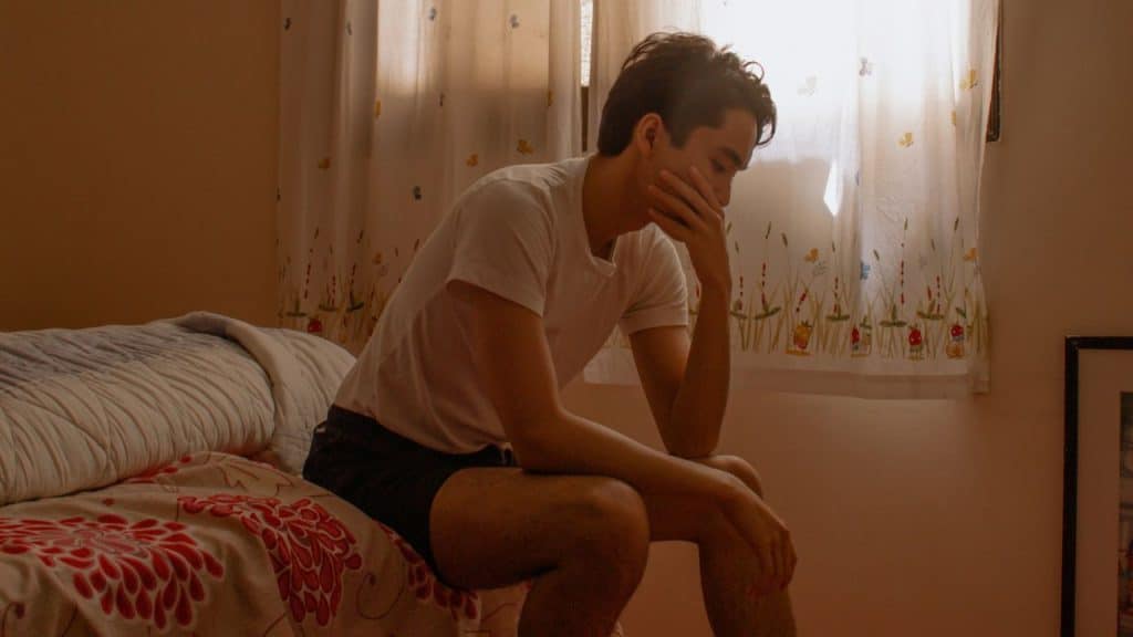 A man sits on the edge of a bed, holding his head in his hand.