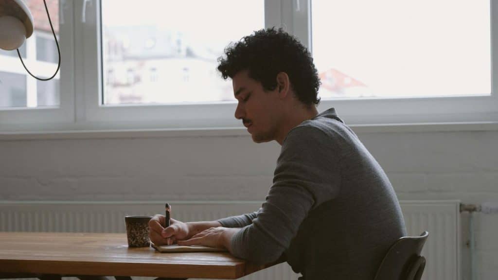 A man with curly hair sits at a wooden table and writes in a notebook.