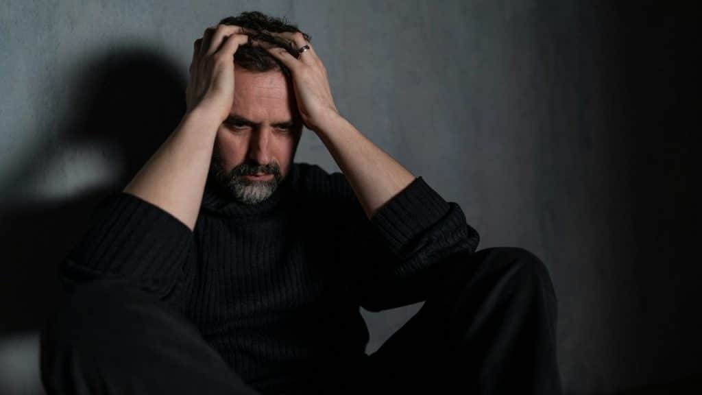 A stressed man sits against a wall, holding his head with both hands.