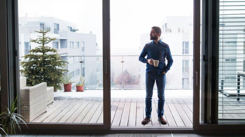 A man stands on a balcony, holding a mug and looking at the view.