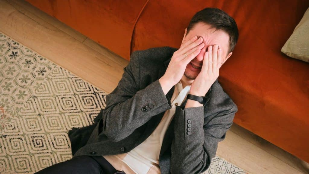 A distraught man in a suit jacket sits on the floor, covering his face.