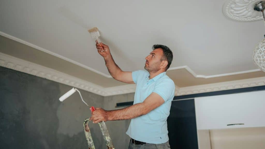 A man on a ladder paints a ceiling with a brush and a paint roller.