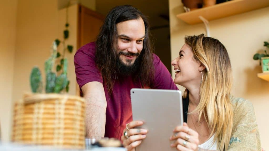 A long-haired man smiles at a laughing woman as they look at a tablet.