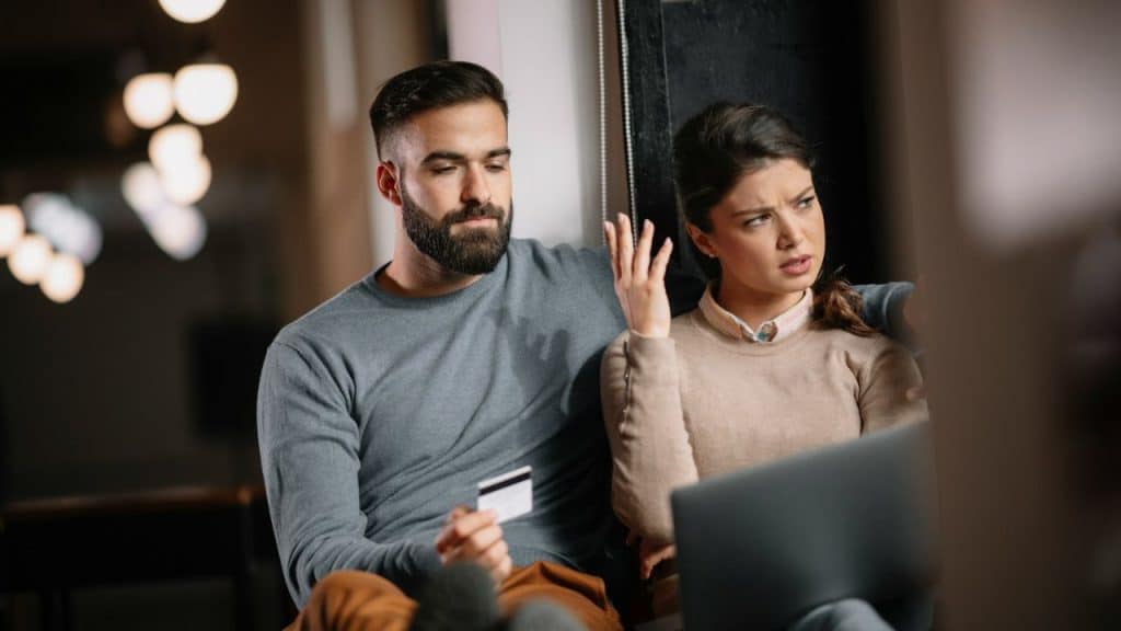 A couple sits together as the man holds a credit card and the woman looks annoyed.