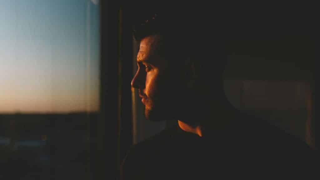 A thoughtful man in profile is lit by sunlight while looking out a window.