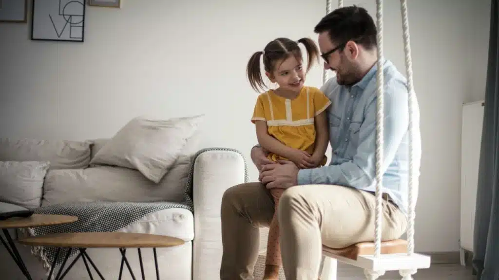 A smiling father sits with his young daughter on a swing inside their home.