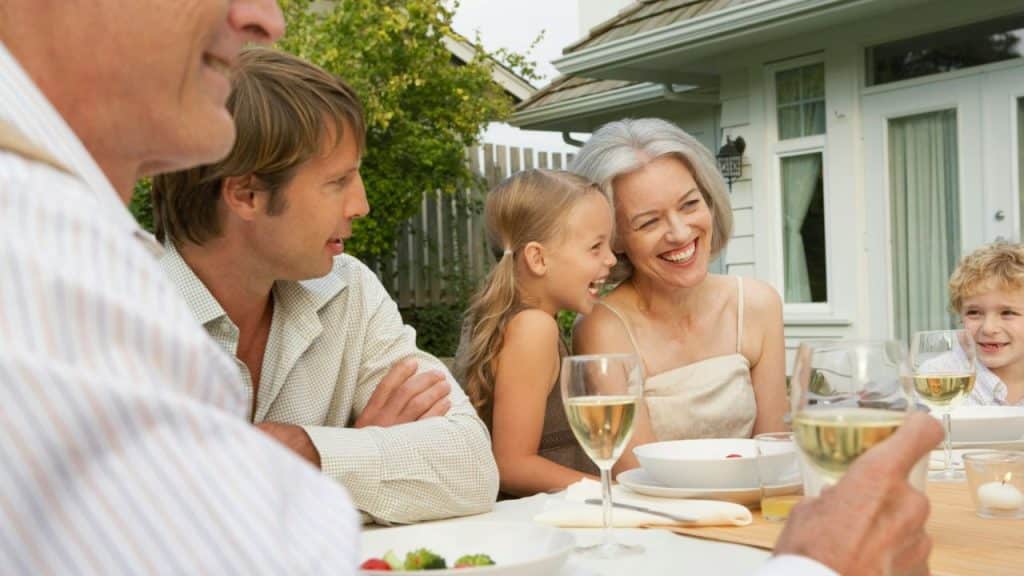 A multigenerational family laughs together while eating and drinking at an outdoor table.