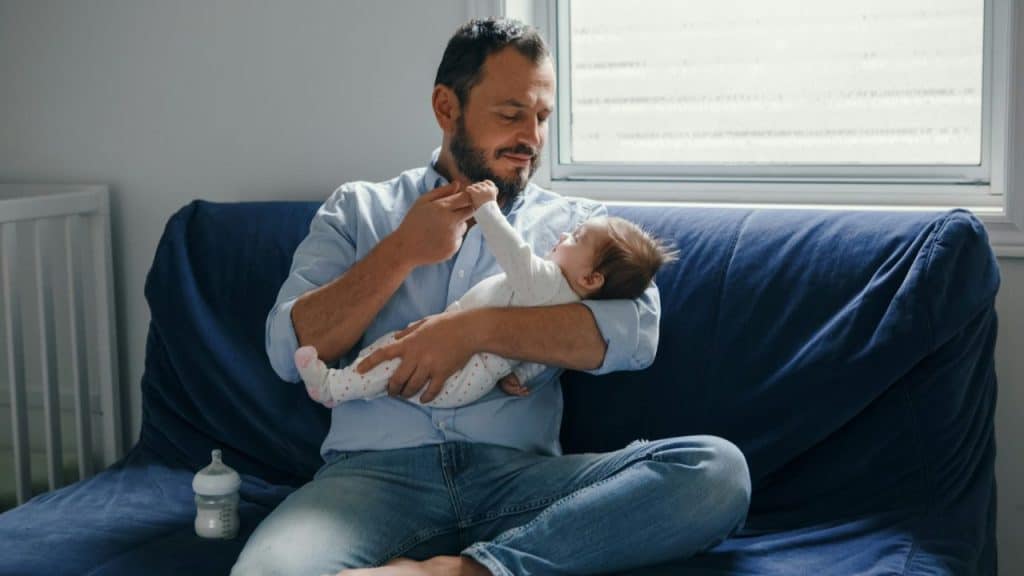 A bearded father in jeans and a blue shirt holds a baby on a blue couch.
