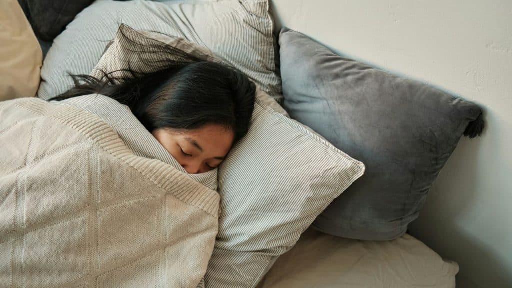 A person with dark hair is sleeping peacefully, covered in a blanket with pillows.