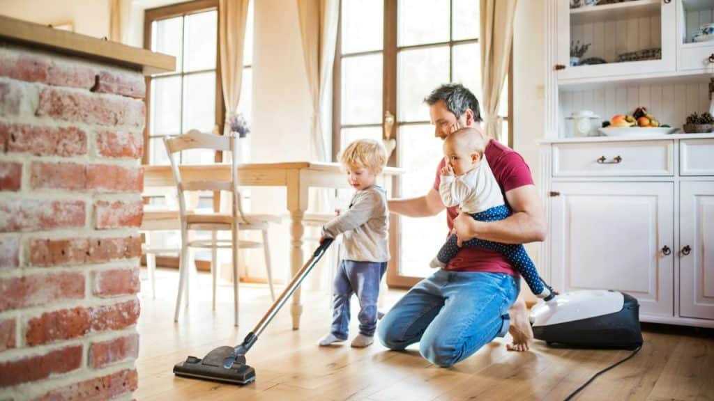 A dad vacuums a wood floor with his two young children at home.
