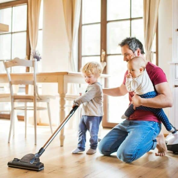 A dad vacuums a wood floor with his two young children at home.