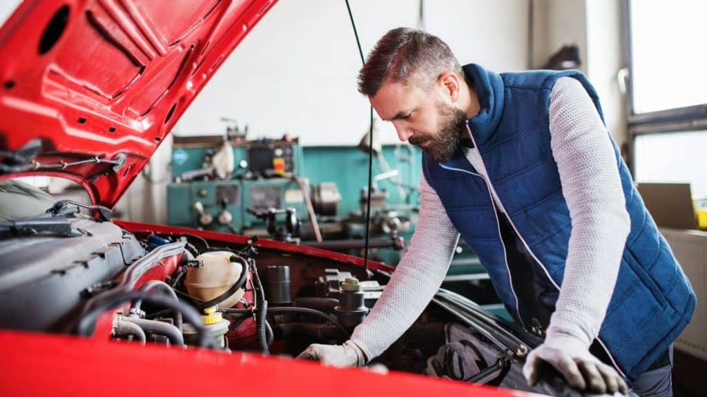 A focused mechanic in a blue vest works on a red car's engine.