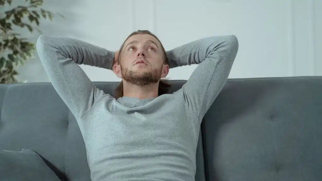 A relaxed man with a beard sits on a couch with his hands behind his head.