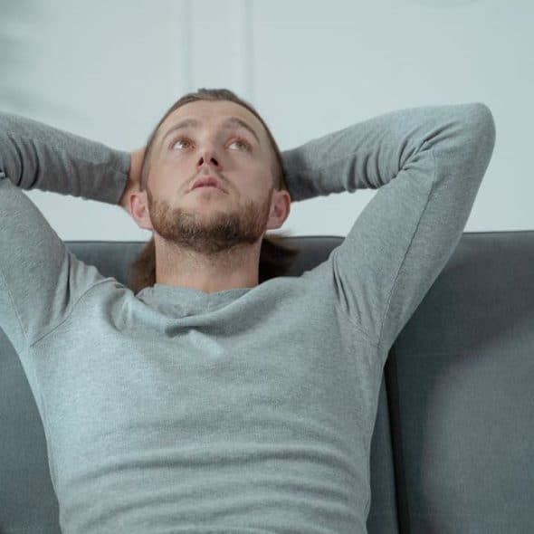 A relaxed man with a beard sits on a couch with his hands behind his head.