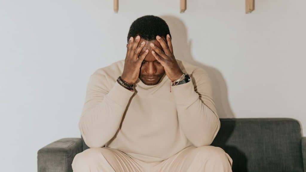 A worried Black man sits on a couch with his head in his hands.