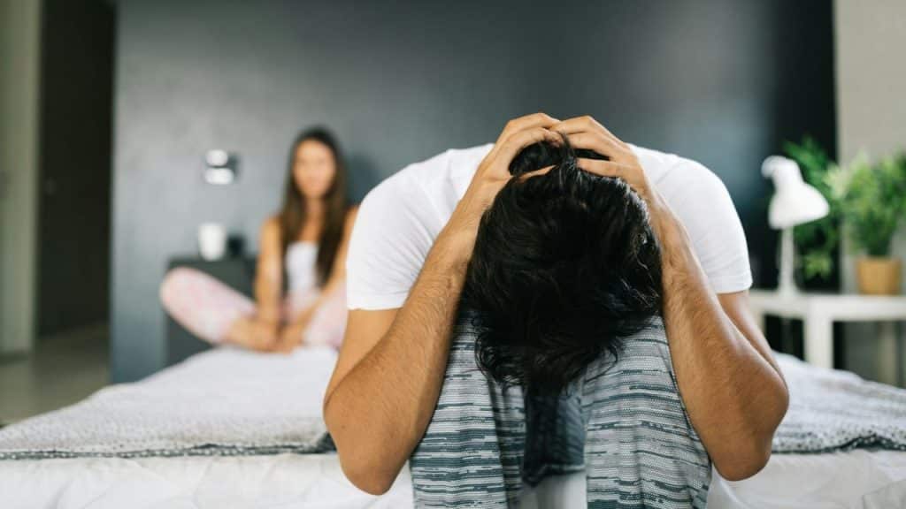 A frustrated man sits with his head in his hands as a woman sits on a bed behind him.