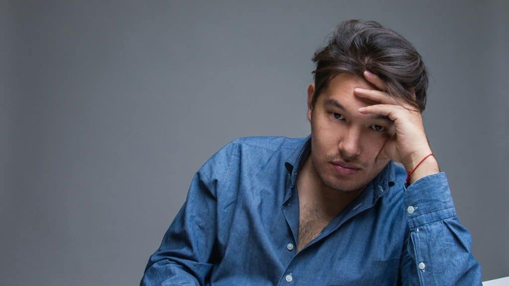 A somber-looking man with long hair rests his chin on his hand.