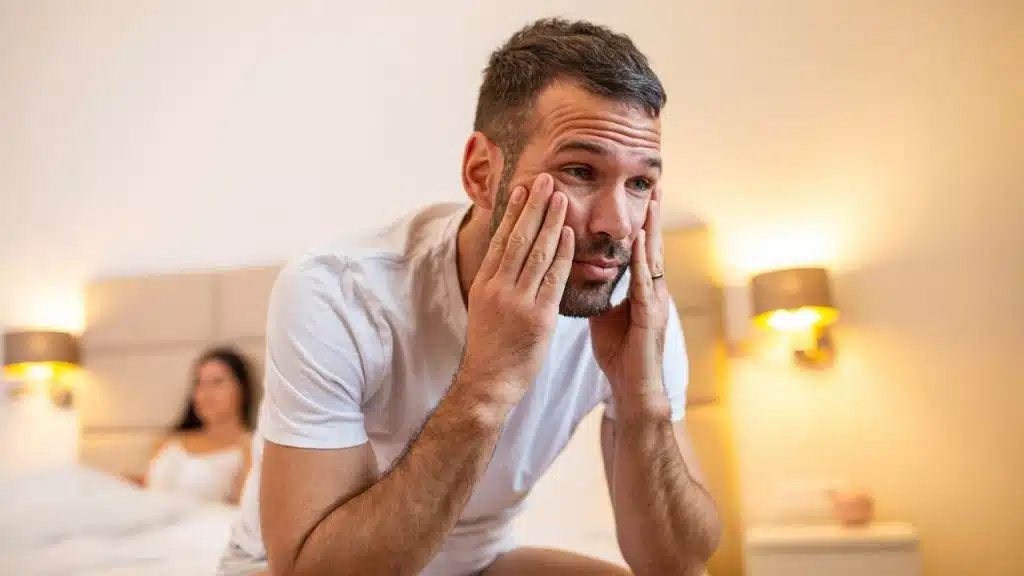 A frustrated man sits on the edge of a bed with his face in his hands, a woman behind him.