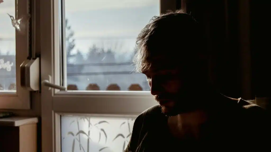 A solemn man in shadow looks down, with light coming from a nearby window.