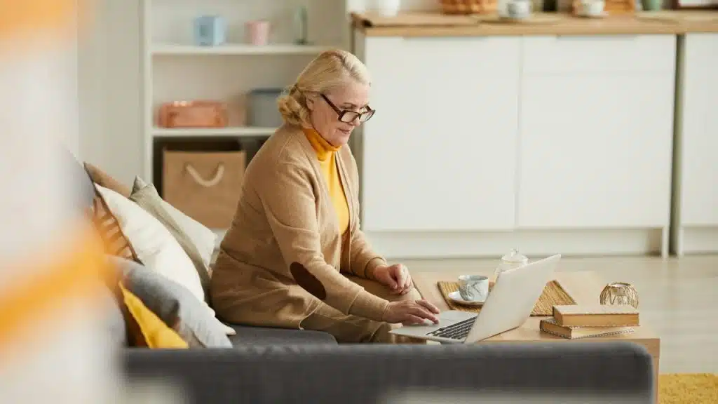 A mature woman in glasses uses a laptop while sitting on a couch in a living room.