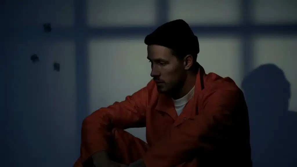 A lonely man wearing an orange jumpsuit and black beanie sits on the floor.