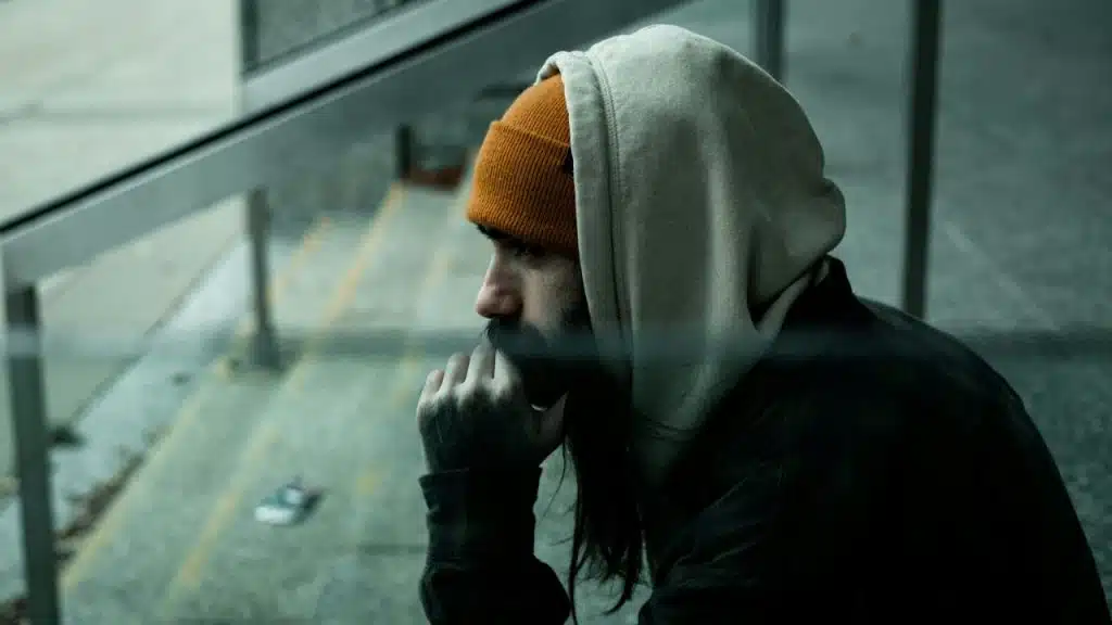 A bearded man in a hoodie and beanie looks contemplatively out a window.