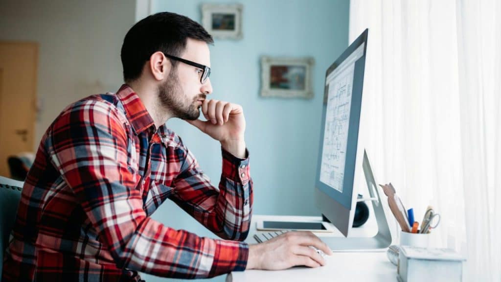 A serious man with glasses and a plaid shirt sits at a desk, looking at a computer screen.