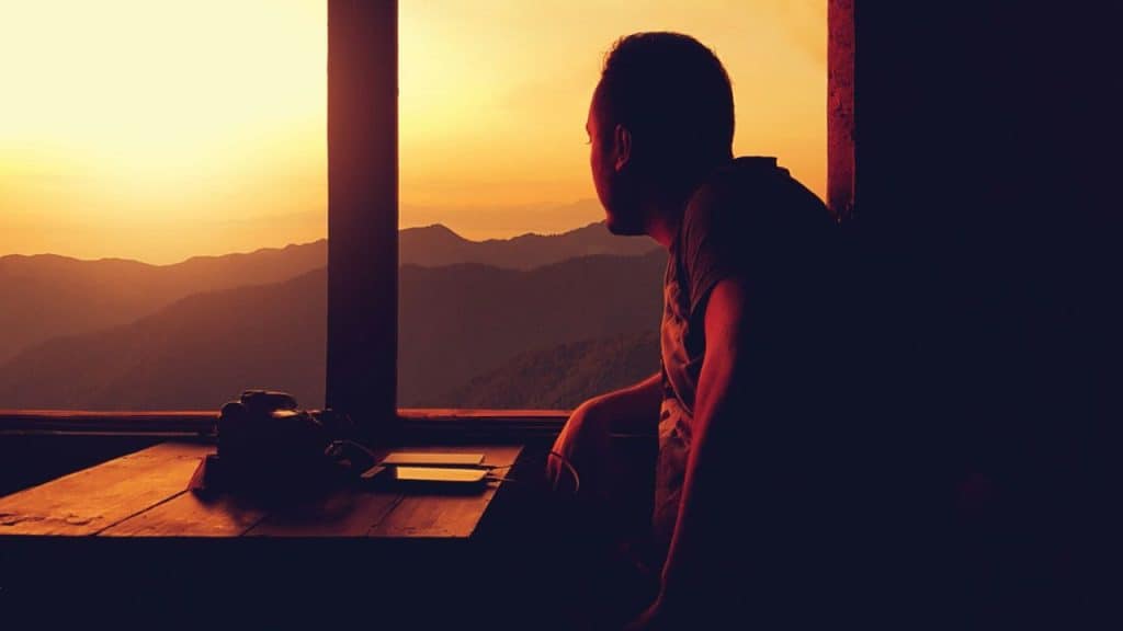 A thoughtful person is silhouetted against a golden sunrise, looking out at mountains.