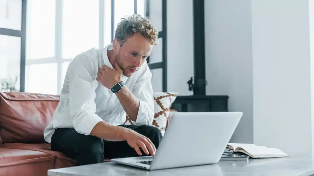 A serious man with blond hair sits on a couch, looking at a laptop.