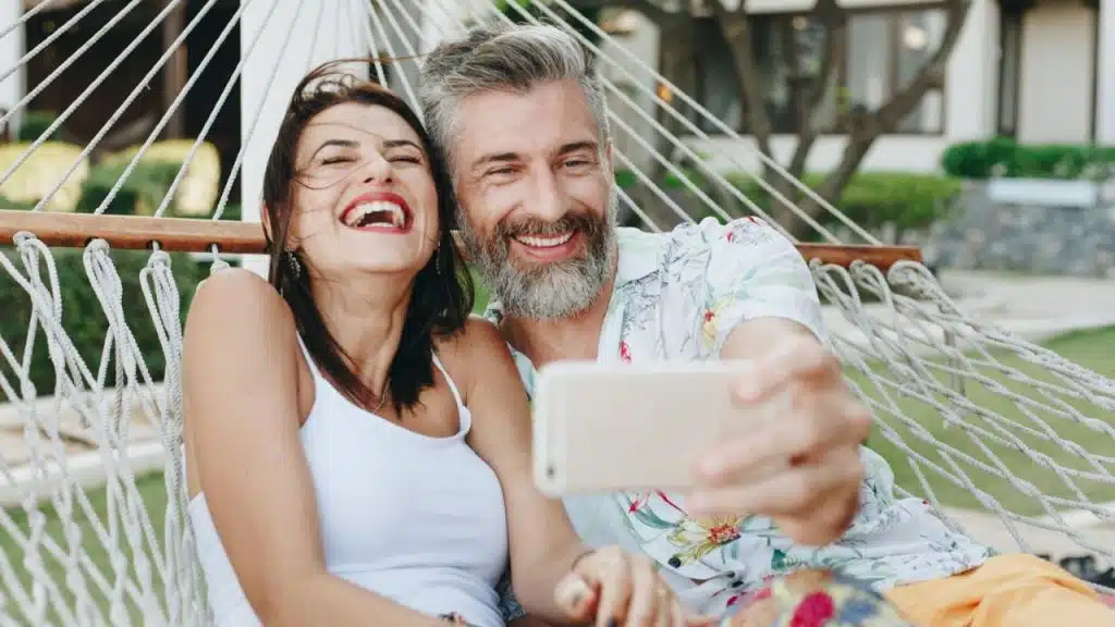 A laughing couple in a hammock takes a selfie with a smartphone.
