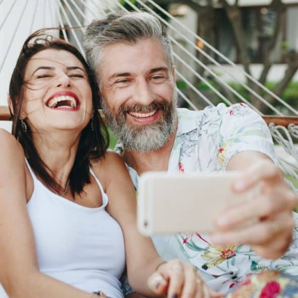 A laughing couple in a hammock takes a selfie with a smartphone.