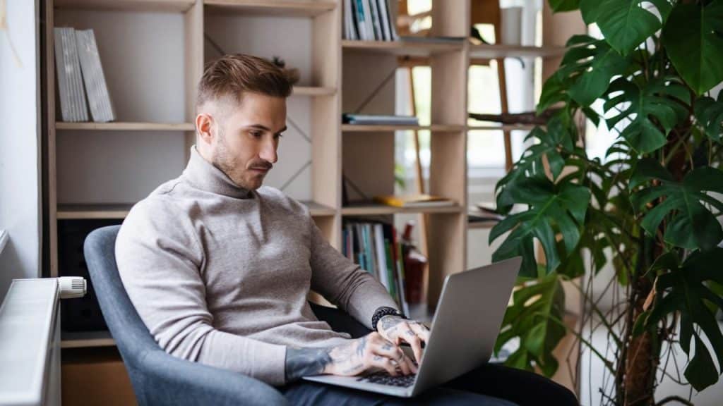 A stylish man with tattoos on his arms sits in a chair, typing on a laptop.