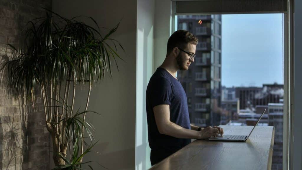 A focused man in glasses stands at a counter, typing on a laptop with a plant beside him.