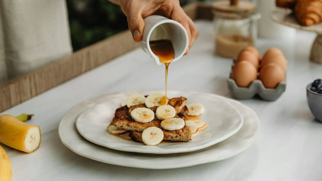 A close-up shot of a person pouring syrup over French toast with banana slices.