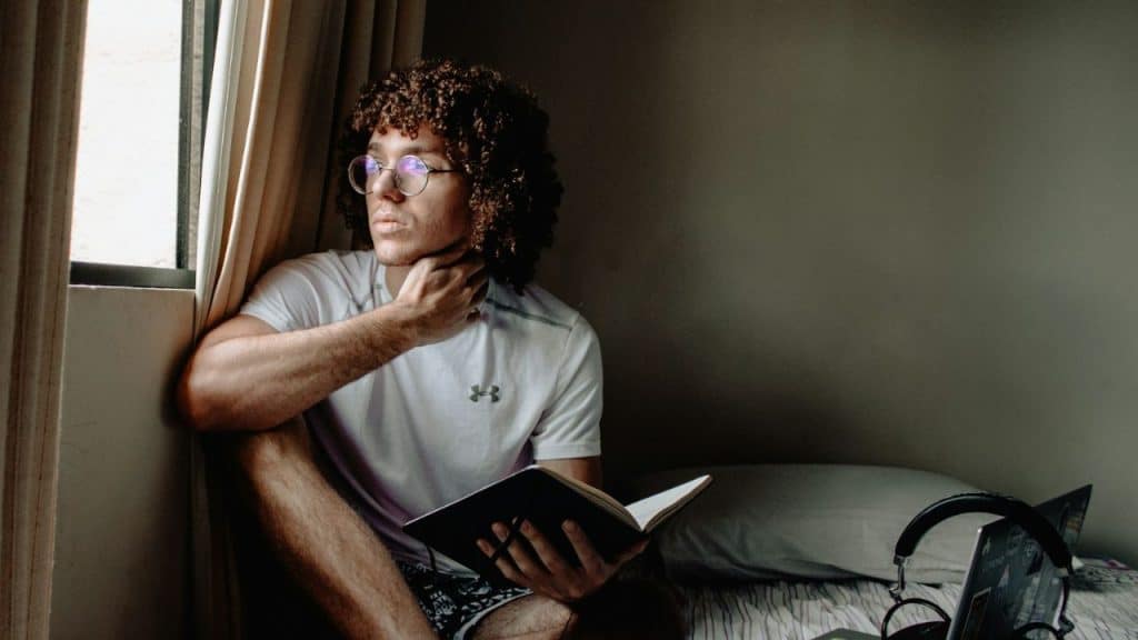 A contemplative man with curly hair and glasses sits near a window, holding a book.