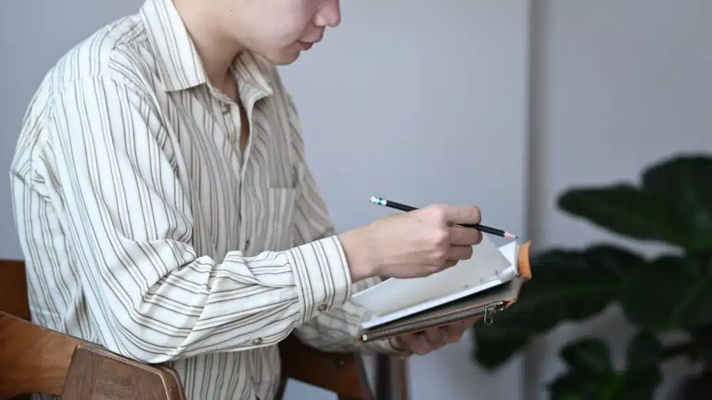A focused person in a striped shirt sits, holding a pencil and a notebook.