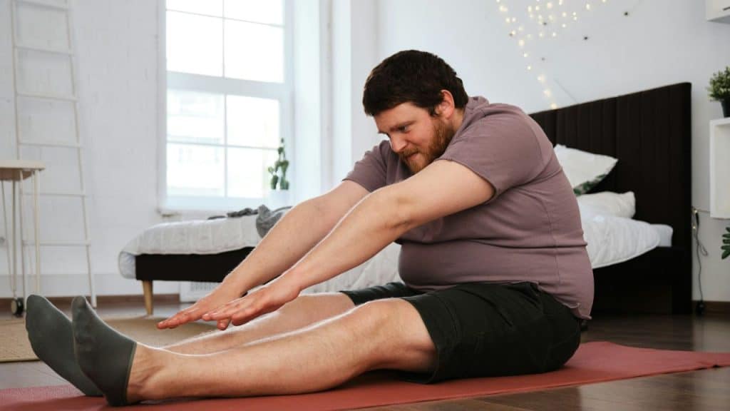 A smiling, bearded man in a t-shirt and shorts stretches on a yoga mat.