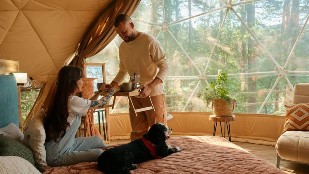 A man brings a woman breakfast in bed inside a geodesic dome, with a dog nearby.