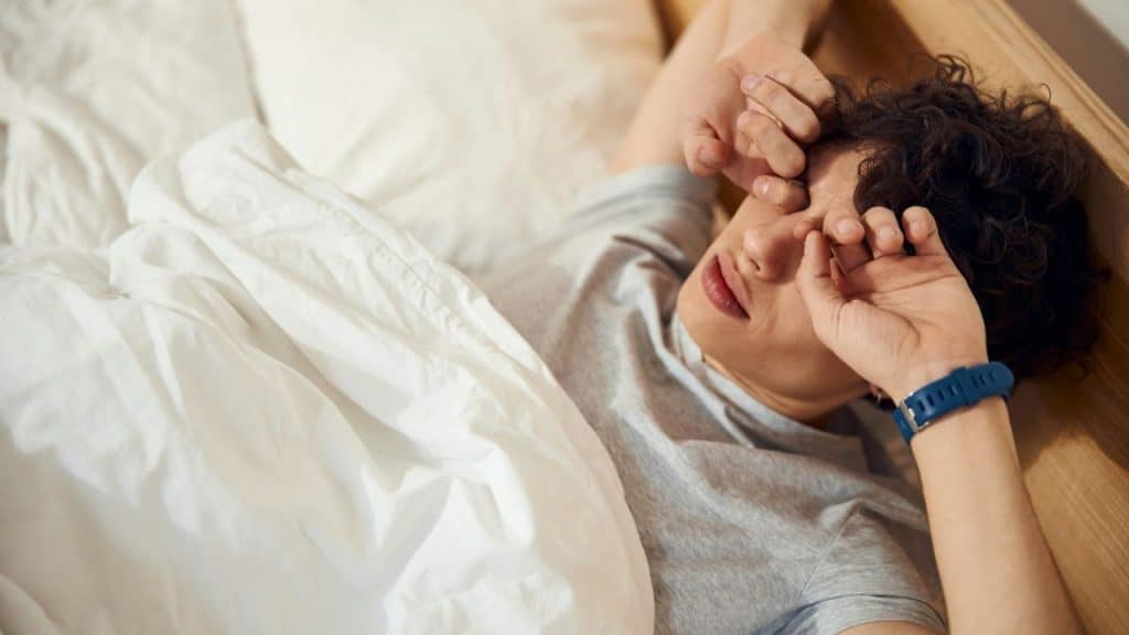 A man with curly hair rubs his eyes while lying in bed.