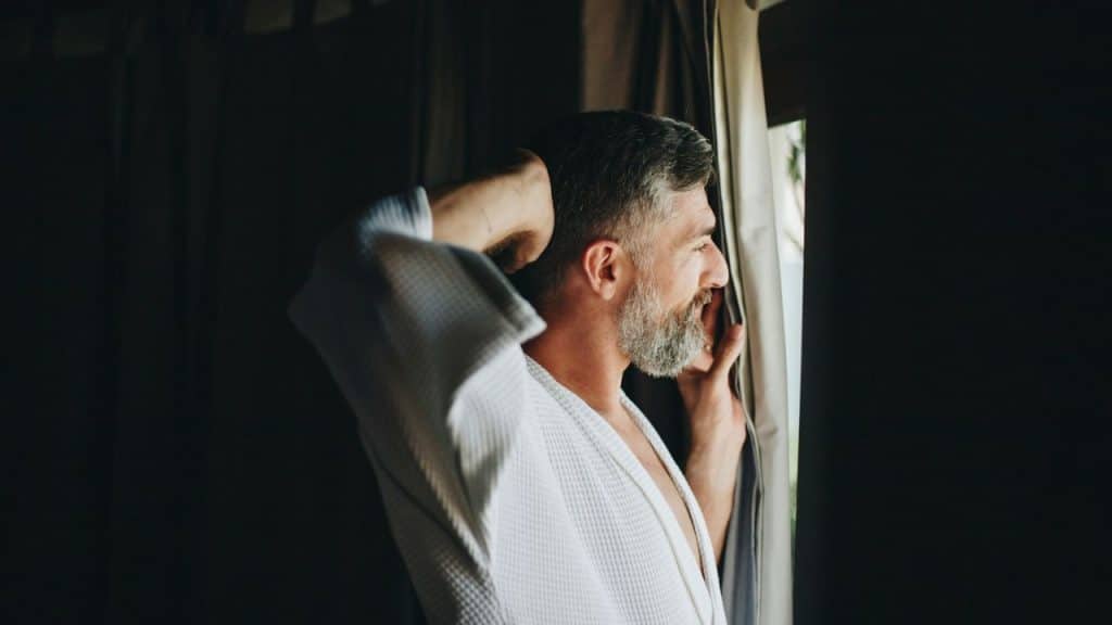 A bearded man in a white robe stands by a window, smiling.