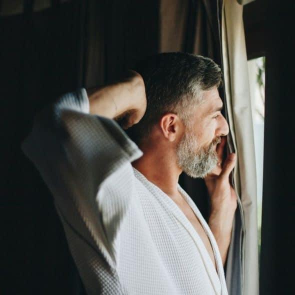 A bearded man in a white robe stands by a window, smiling.