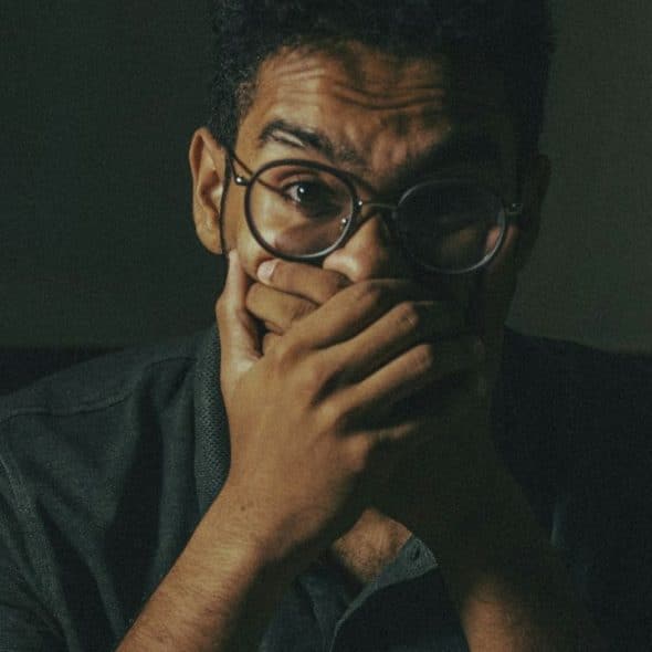 A startled-looking man with glasses and curly hair covers his mouth with his hands.